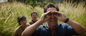 Three people stand in tall grass; the person in front is looking through binoculars, while the two behind watch attentively.