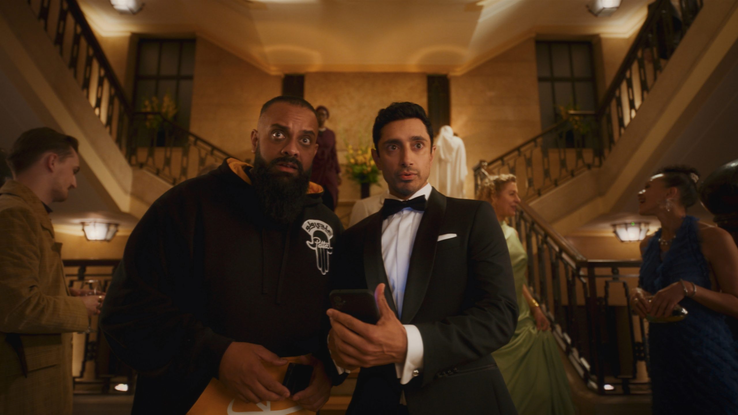 Two men, one in a hoodie and one in a tuxedo, stand in a grand lobby looking surprised, surrounded by other people in formal attire.