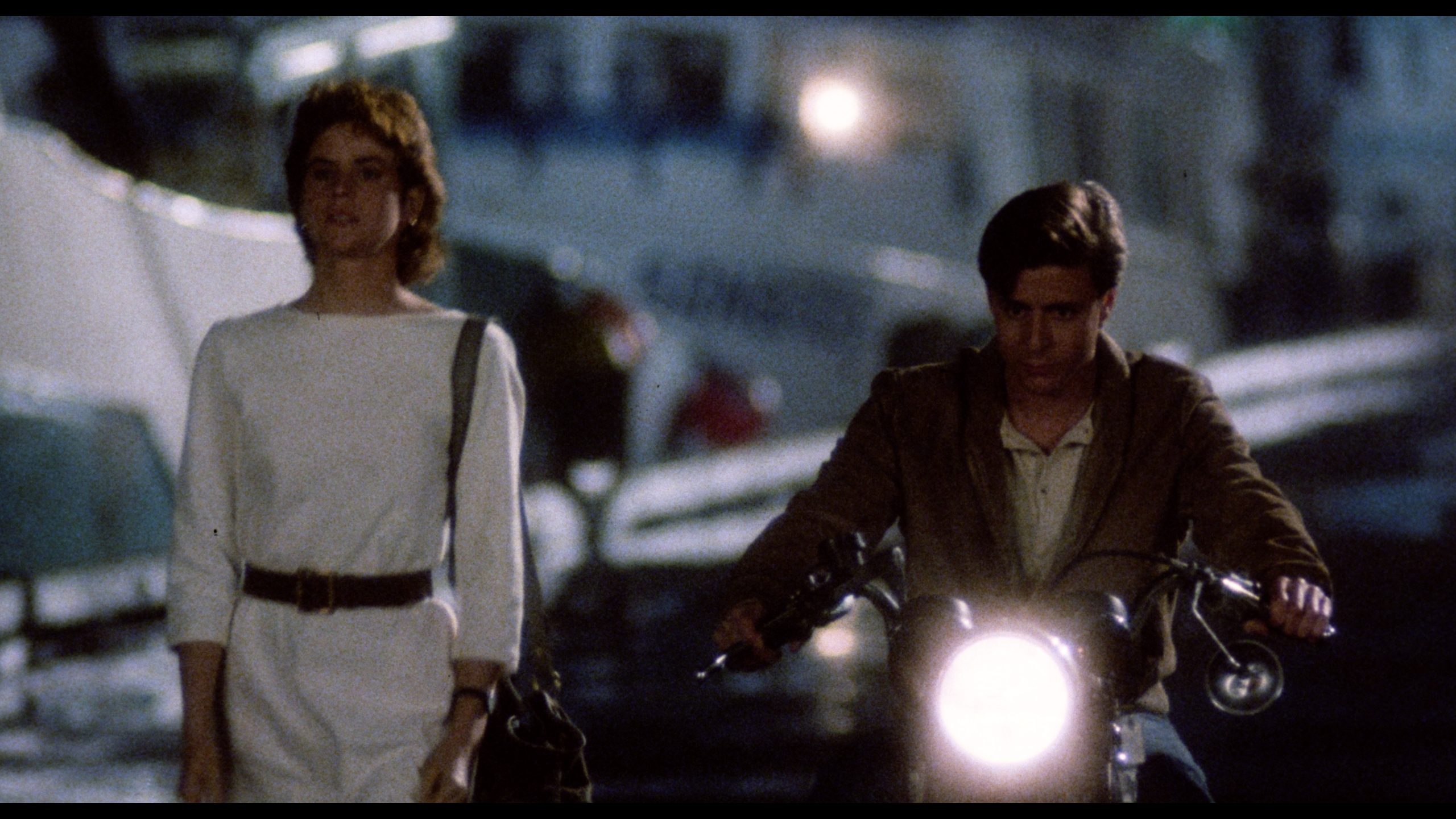 A woman in a white dress walks beside a man riding a motorcycle at night; the background is slightly out of focus.