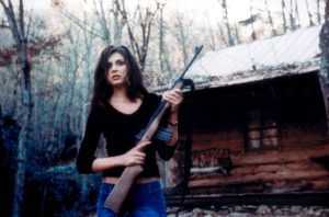 A woman in a black top and jeans stands outdoors holding a rifle in front of a rustic log cabin surrounded by trees.