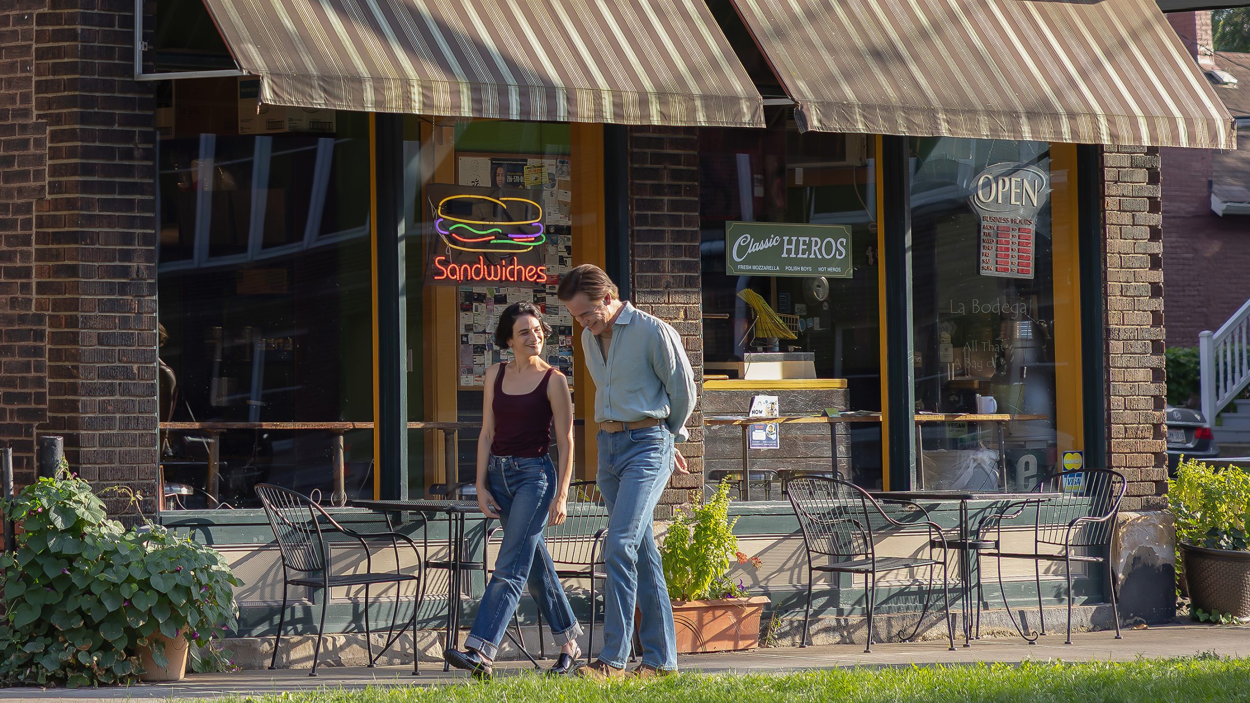 Two people walk past outdoor tables in front of a brick sandwich shop with striped awnings and neon signs in the window.