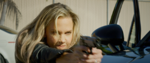 A woman with blonde hair aims a handgun while crouching beside a black car, appearing focused and intense.