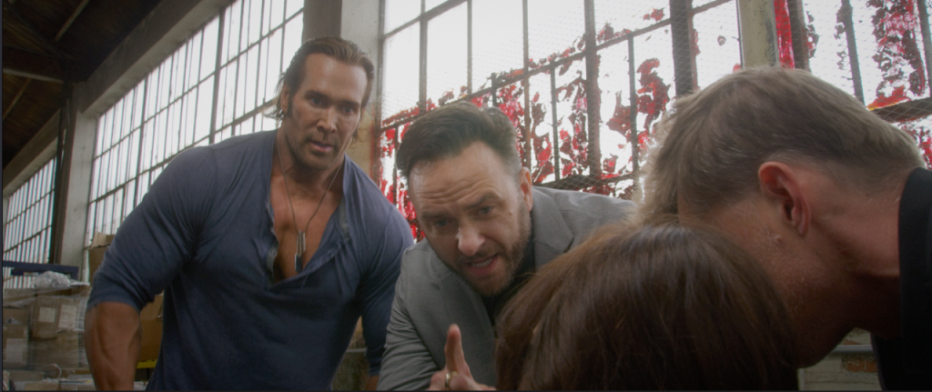 Three men are gathered around someone sitting down in a warehouse with large windows and a blood-splattered wall in the background.