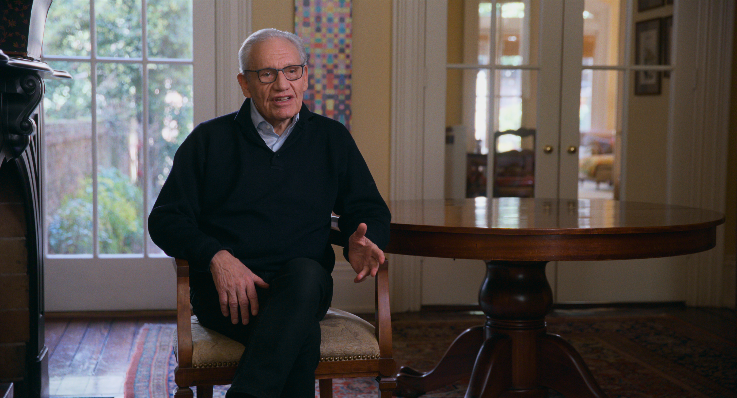 An older man wearing glasses and a dark sweater sits in a wooden chair, speaking, with a round table and large windows in the background.
