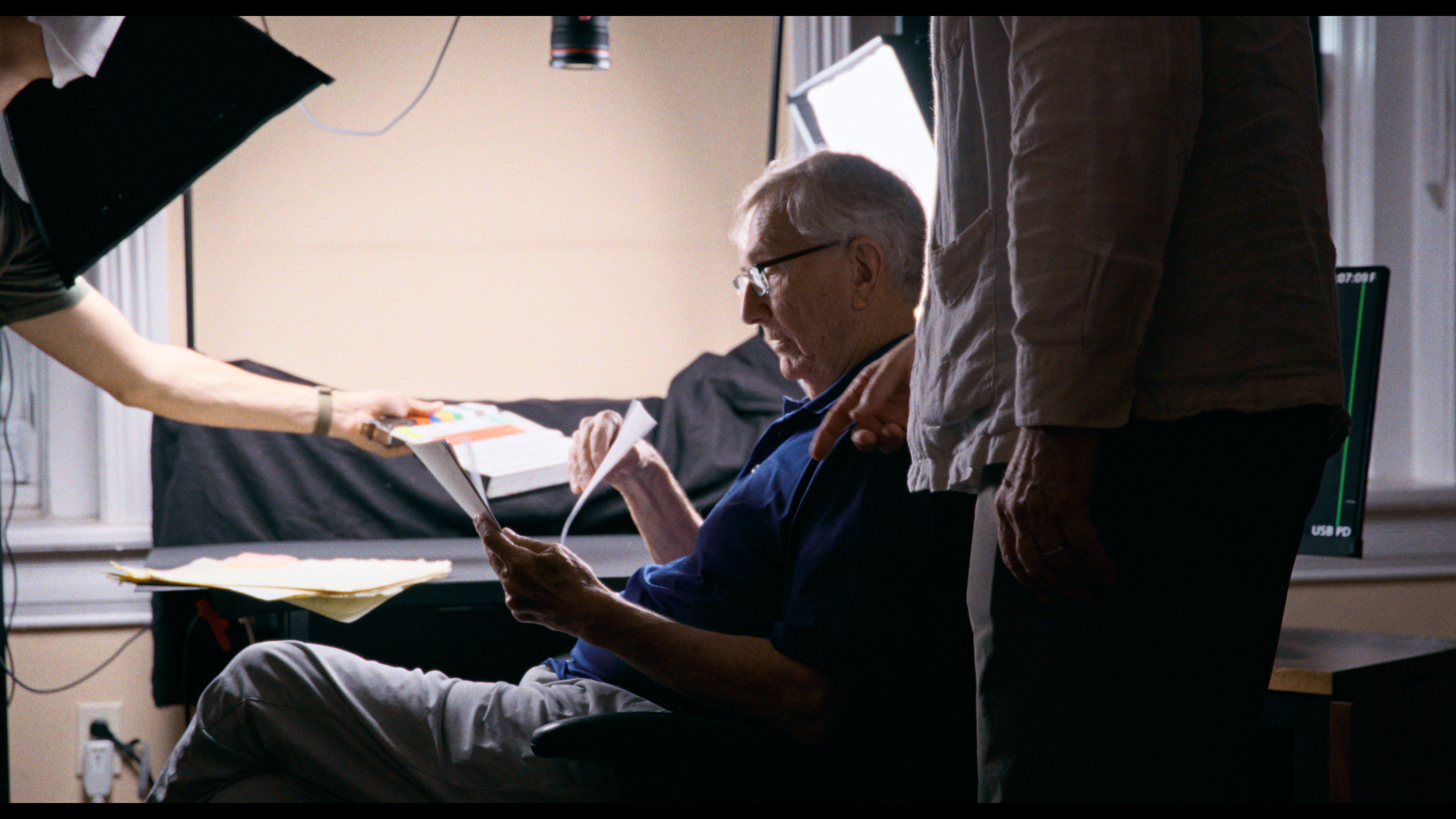 An older man sits in a chair reading papers while another person stands beside him and a third person hands him a book in a room with studio lights and filming equipment.