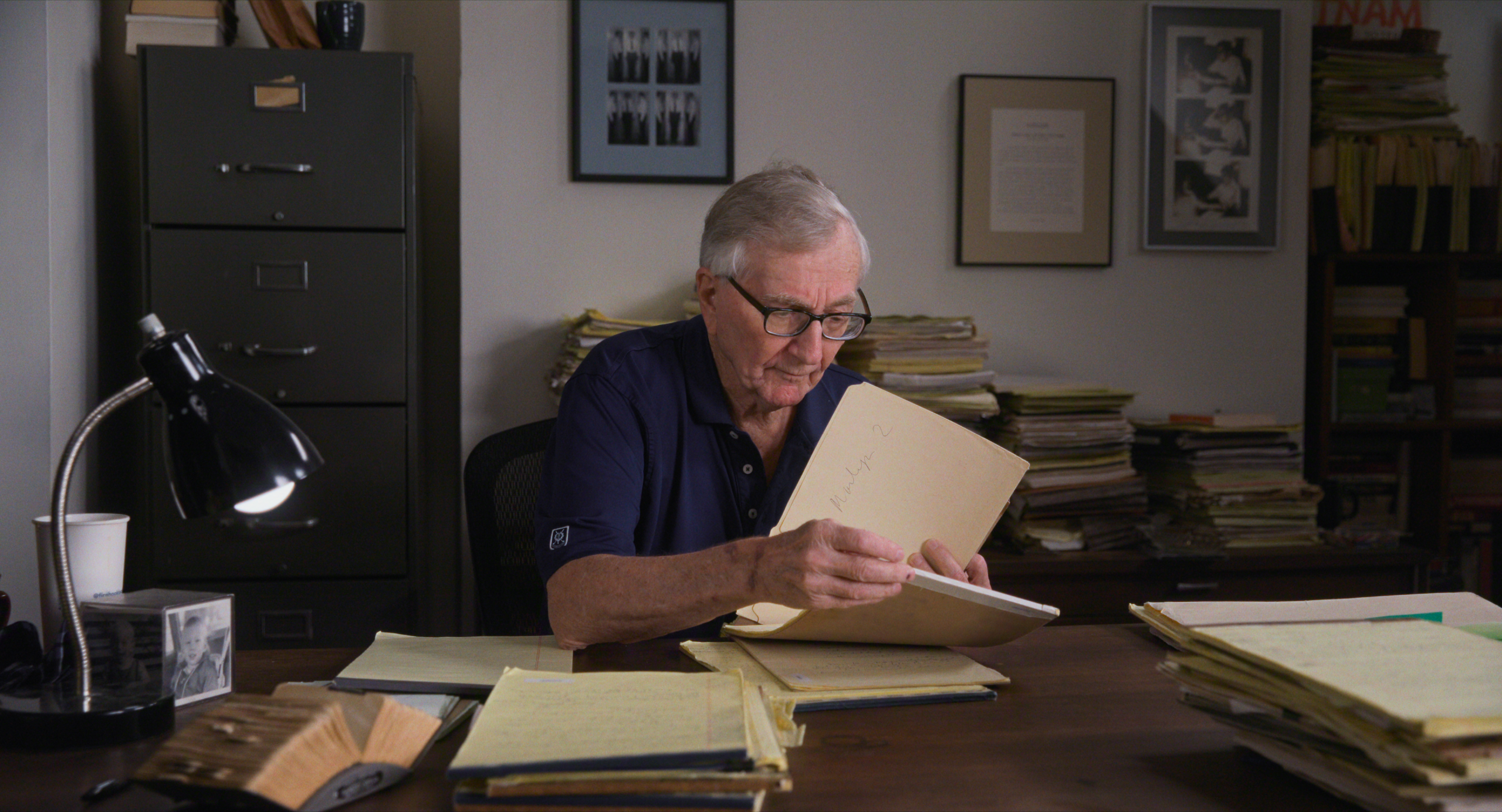 An older man wearing glasses sits at a desk covered with papers, looking through a file folder in an office with stacked documents and framed pictures on the wall.