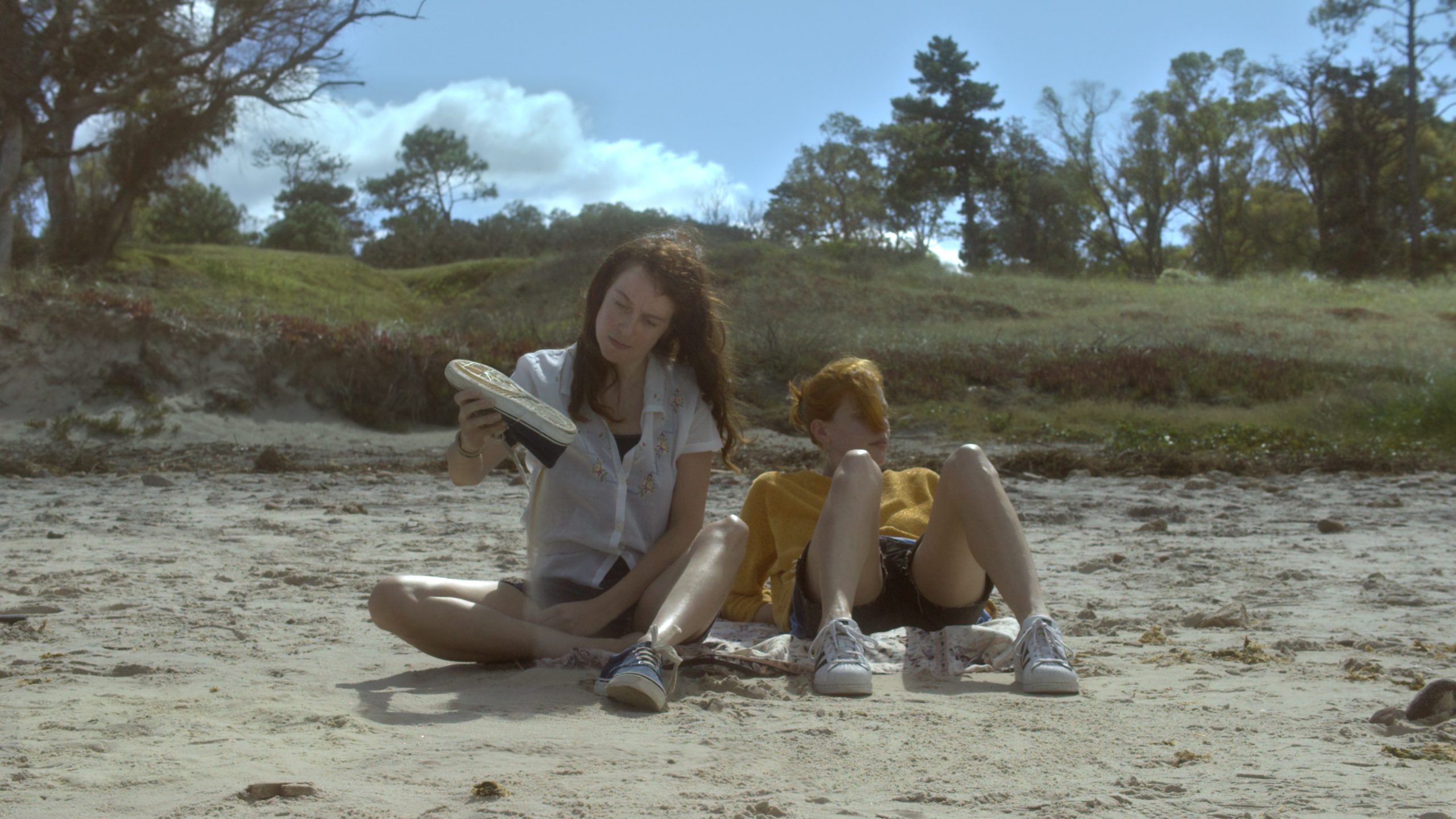 Two people sit on a sandy beach, one holding a shoe and the other reclining with knees up. Trees and grass are visible in the background under a partly cloudy sky.