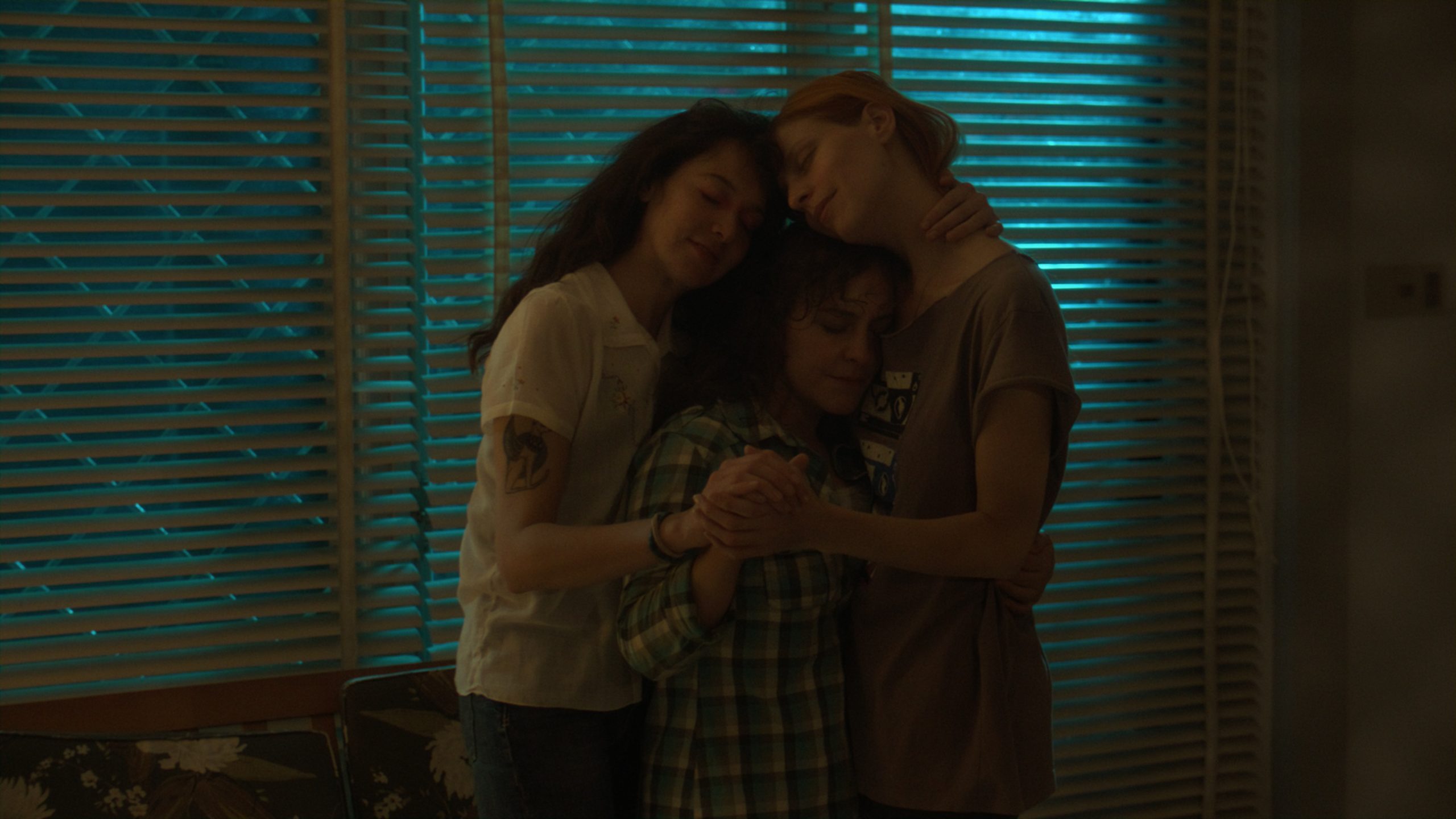 Three people stand close together, eyes closed, sharing an embrace in a dimly lit room with closed blinds and blue light filtering through.