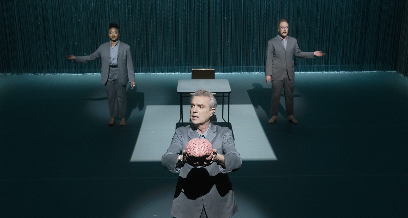 Three people in gray suits perform on stage; the person in front holds a model of a brain, with a desk and laptop behind them under dramatic lighting.