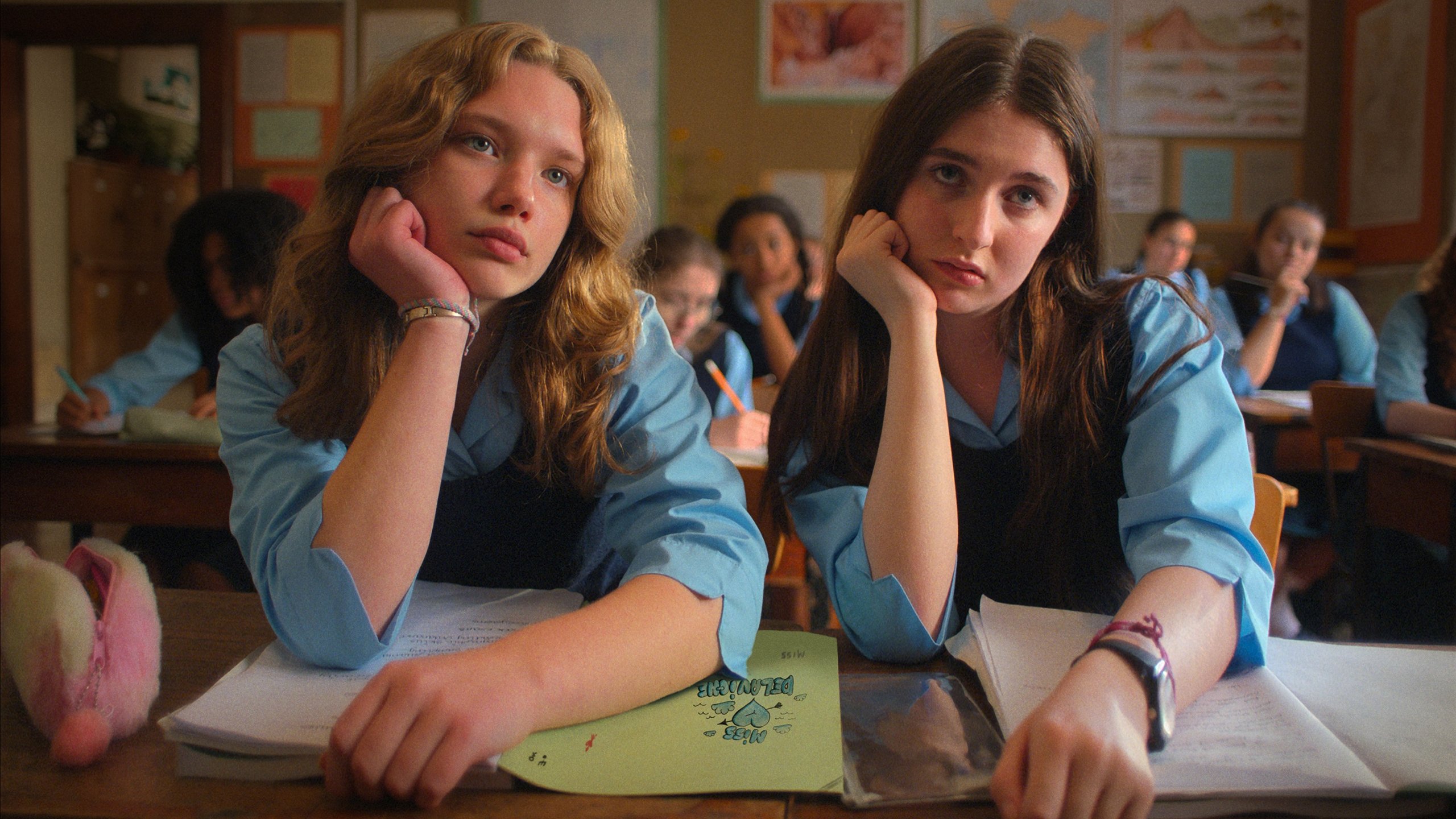 Two teenage girls in school uniforms sit at desks in a classroom, resting their heads on their hands and looking bored, with open notebooks in front of them.