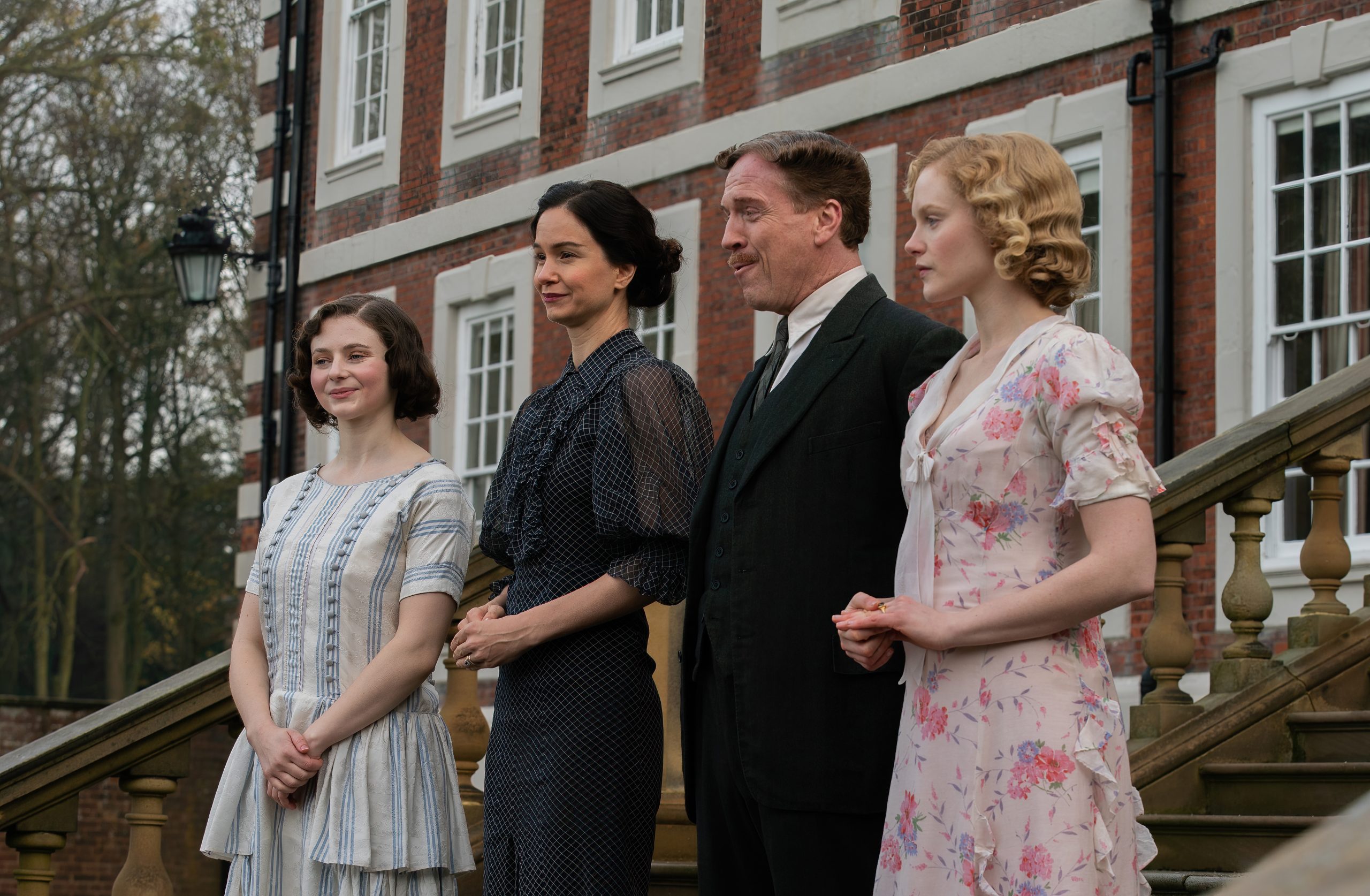 Four people in period clothing stand side by side outside a large brick building with white windows and a wooden staircase.