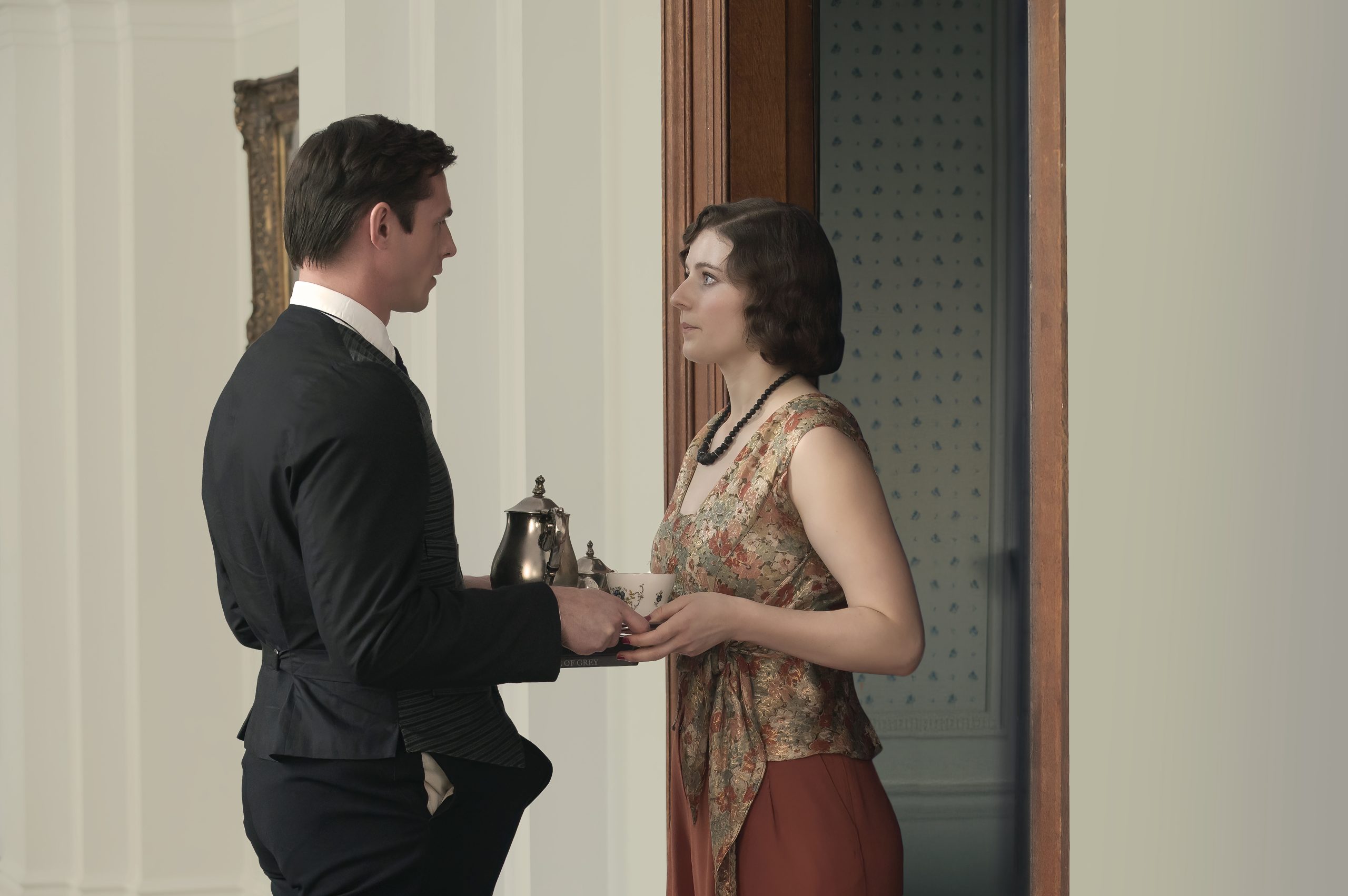 A man in formal attire hands a tray with a teapot and cup to a woman in patterned clothing as they stand near a doorway inside a building.