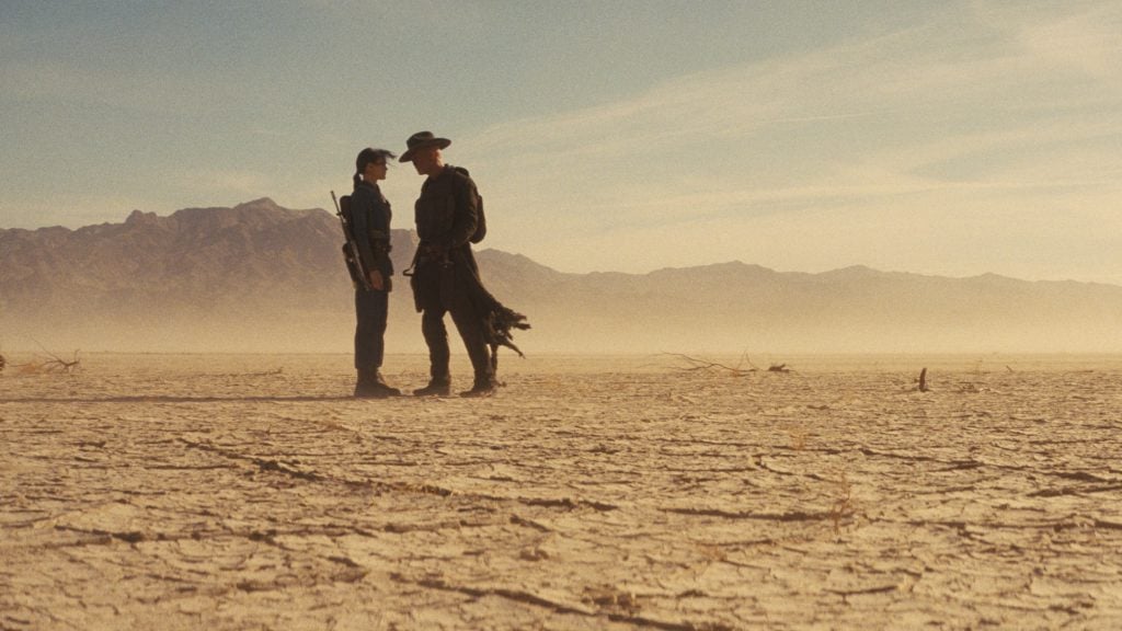 Two people stand facing each other in a barren desert landscape with mountains in the background under a clear sky.