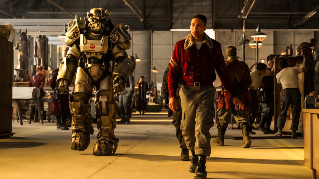 A person in a red jacket walks next to someone in a large armored suit inside a busy industrial warehouse with several people working in the background.