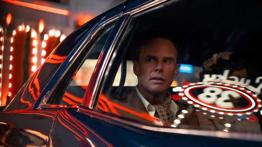 A man in a suit sits in the back seat of a car at night, looking out the window at bright neon lights reflected on the glass.