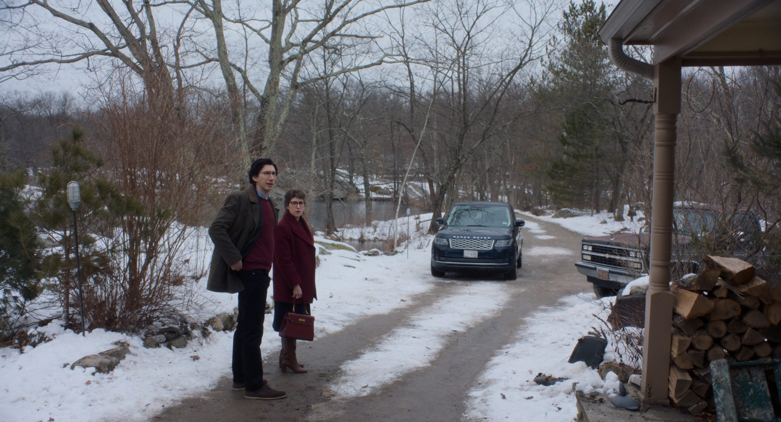 A man and a woman stand on a snowy driveway near a house, with two parked cars and stacked firewood visible. The background shows leafless trees and a pond.