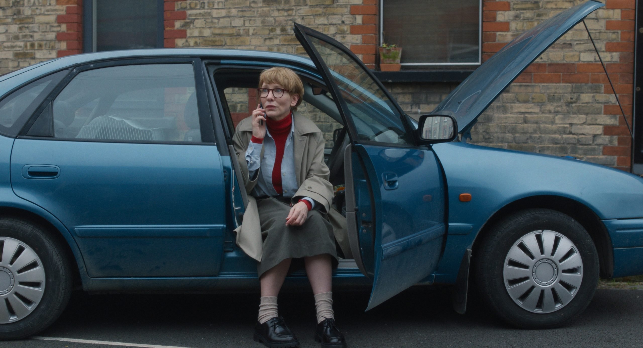 A person wearing glasses and a trench coat sits on the edge of an open car door, smoking a cigarette in a parking lot beside a blue car.