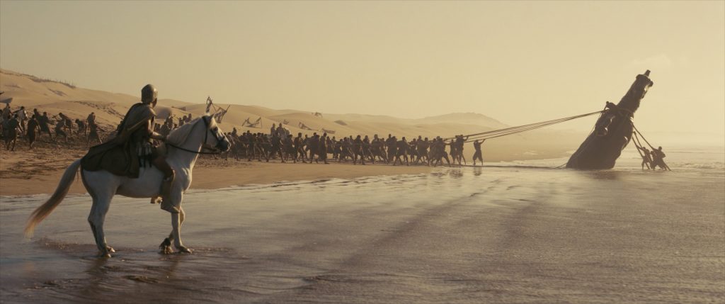 A group of armored soldiers on a beach uses ropes to pull a large, partially buried statue head while a soldier on horseback observes. Sand dunes are visible in the background.