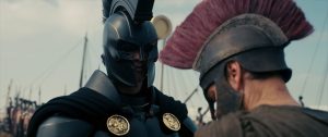 Two men in ancient-style armor and helmets face each other, one in black with gold medallions, the other in bronze with a red crest, with ships in the background.