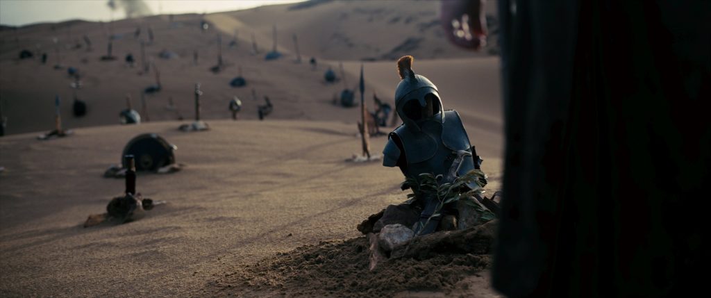 A desert graveyard with scattered helmets, armor, and swords marking graves, with a close-up of a helmet and armor piece in the foreground.
