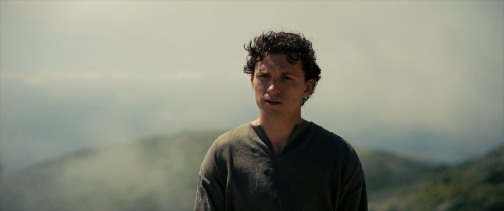 A young man with curly hair stands outdoors in front of a hazy, mountainous background, wearing a loose, dark shirt.