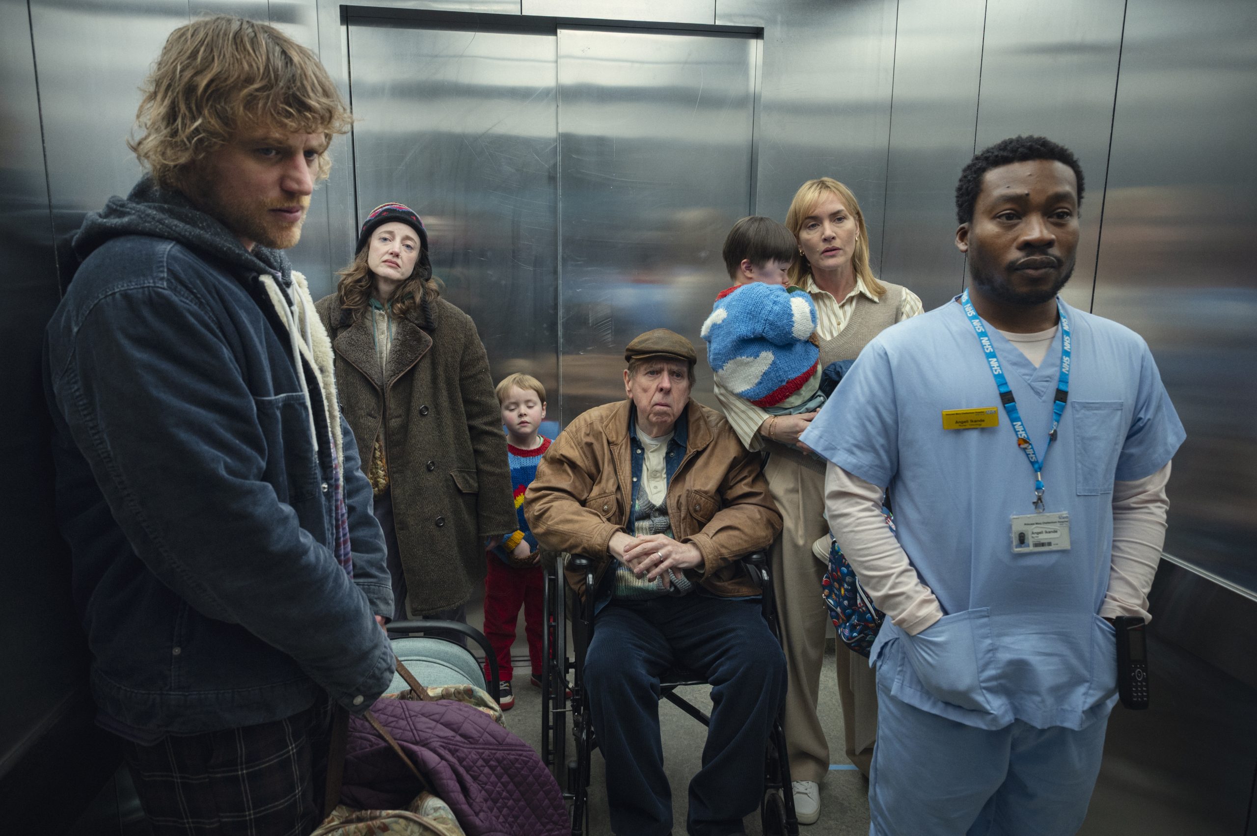 Seven people, including a nurse, a man in a wheelchair, a woman holding a child, and others, stand in a crowded elevator with reflective metal walls.