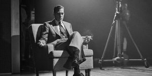 A man in a suit sits in an armchair on a stage, holding a cigarette, with studio lighting and camera equipment in the background. The image is in black and white.
