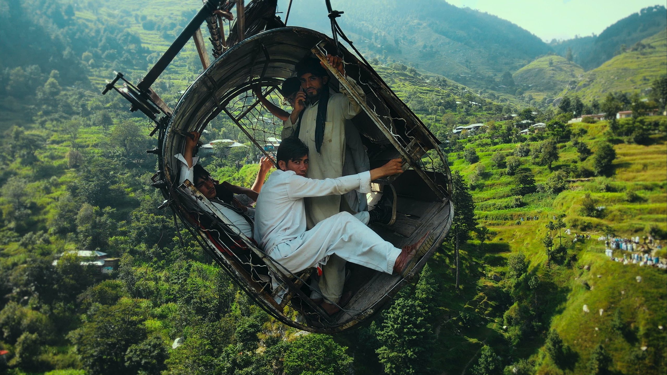 Several people ride in a worn cable car suspended above a lush, green mountainous landscape.