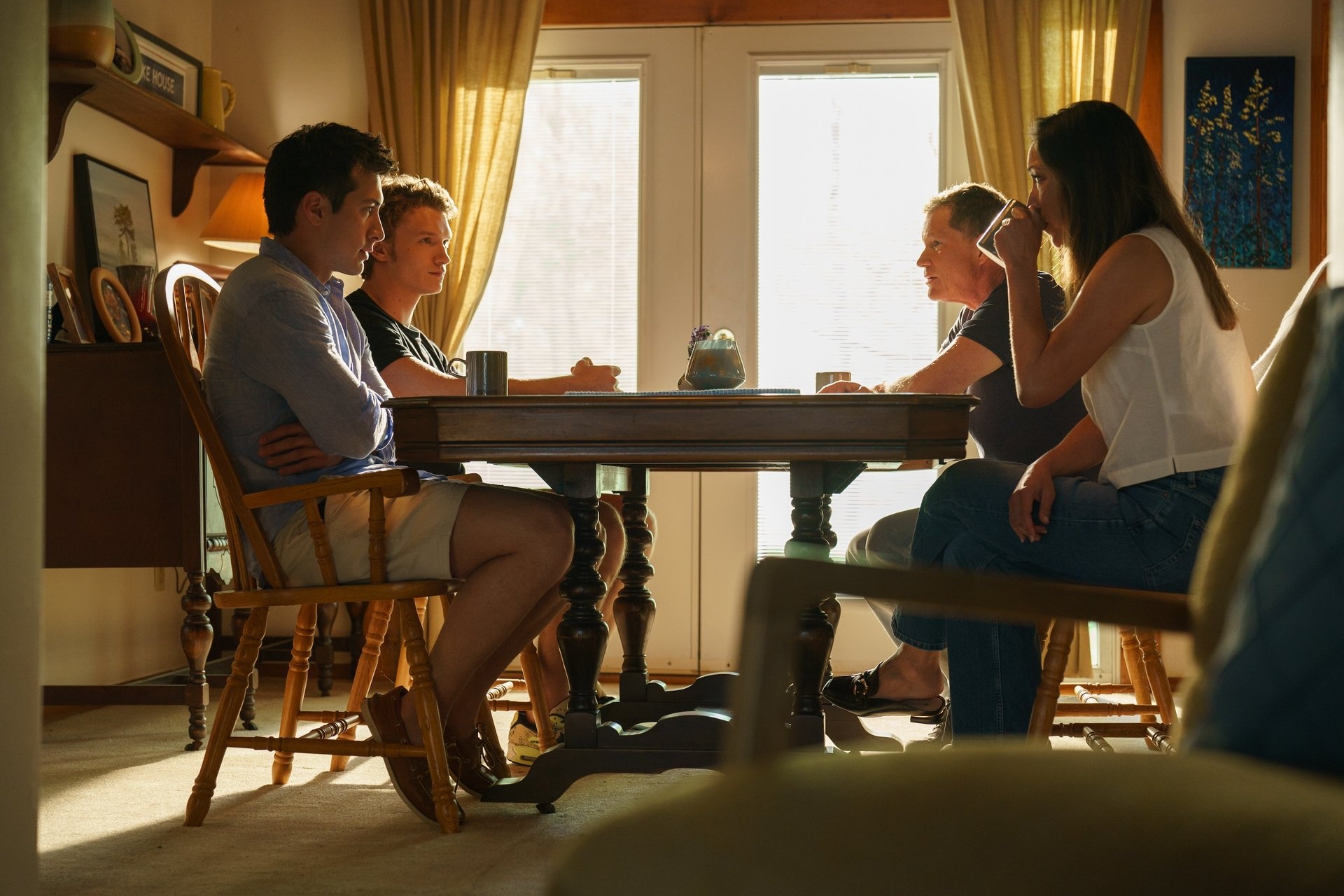 Four people sit around a wooden table in a sunlit room, engaged in conversation. Cups and a teapot are on the table, and sunlight streams through glass doors behind them.