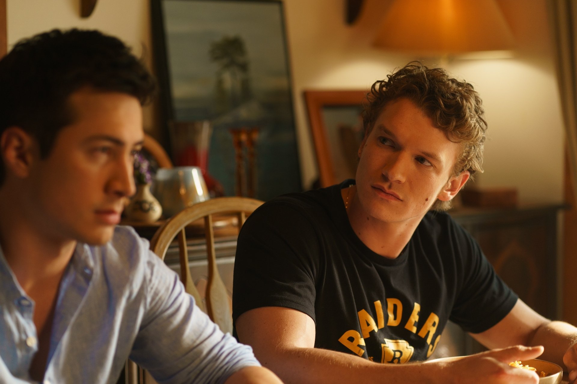 Two young men sit at a wooden table indoors; one looks serious while the other gazes at him with a questioning expression. Framed artwork and household items are visible in the background.