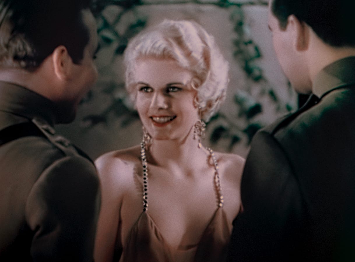 A woman with short blonde hair and a satin dress smiles while talking to two men in military uniforms.
