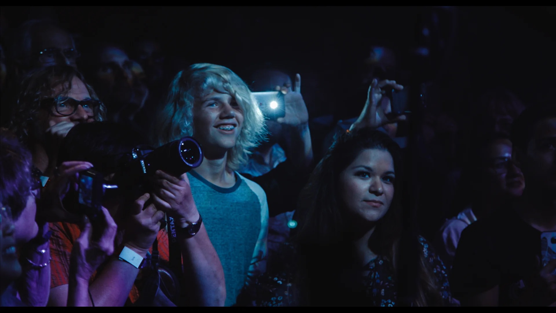 A group of people standing in a dark setting, some holding cameras or phones to take photos, illuminated by stage lights.
