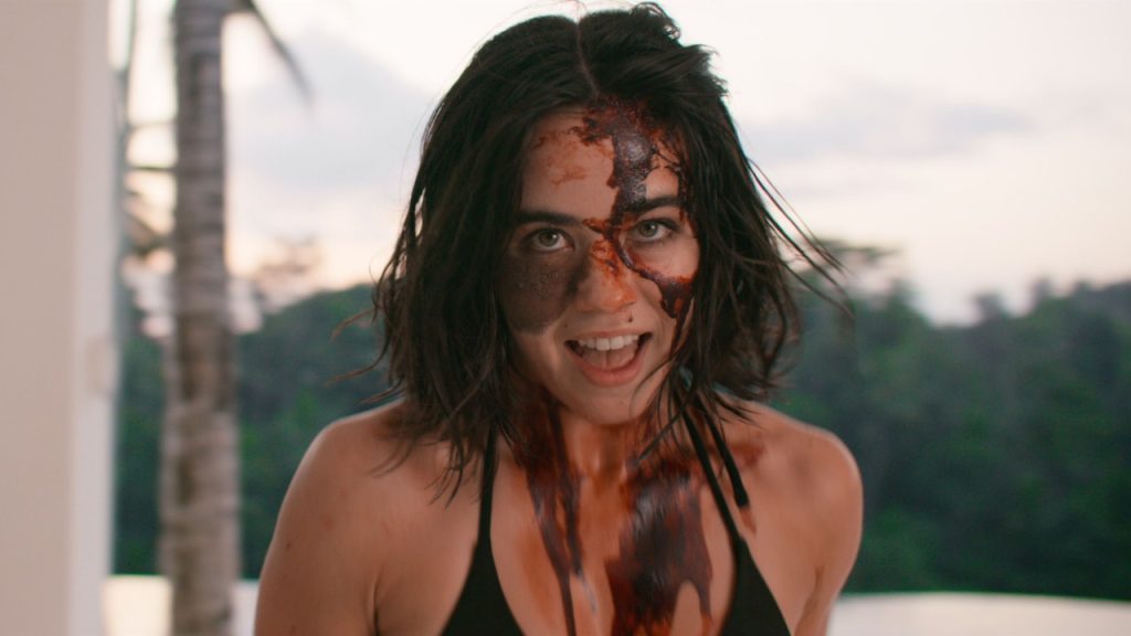 A woman with messy dark hair and a black top stands outdoors, her face and chest smeared with a dark, blood-like substance. She looks directly at the camera with an intense expression.