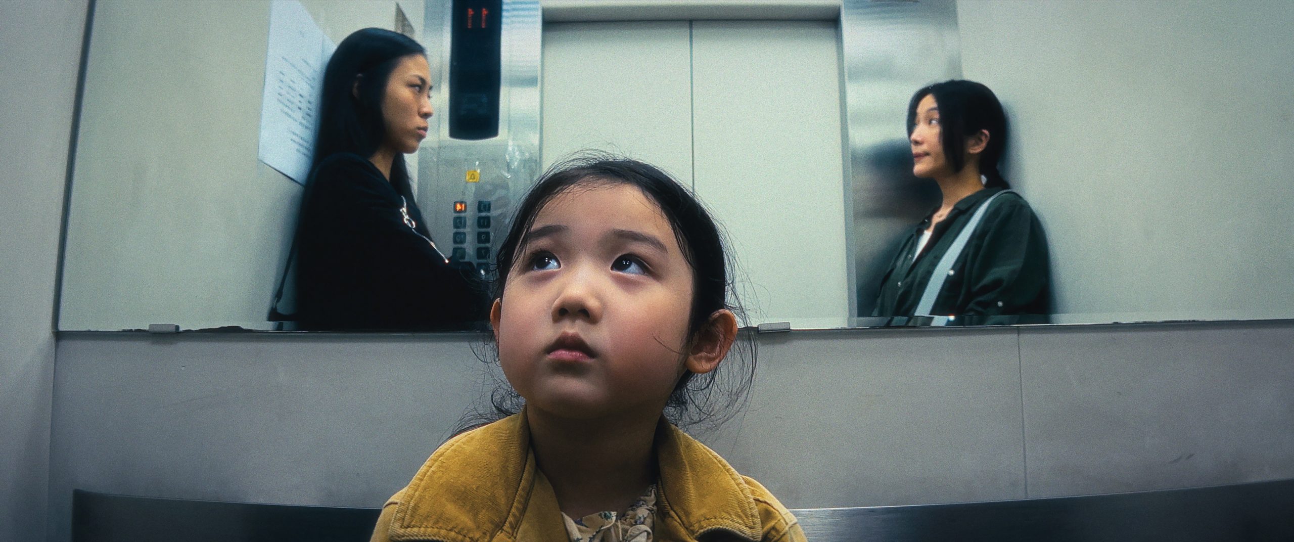A young girl looks up while seated in an elevator, with two women standing on opposite sides staring at each other in the background.