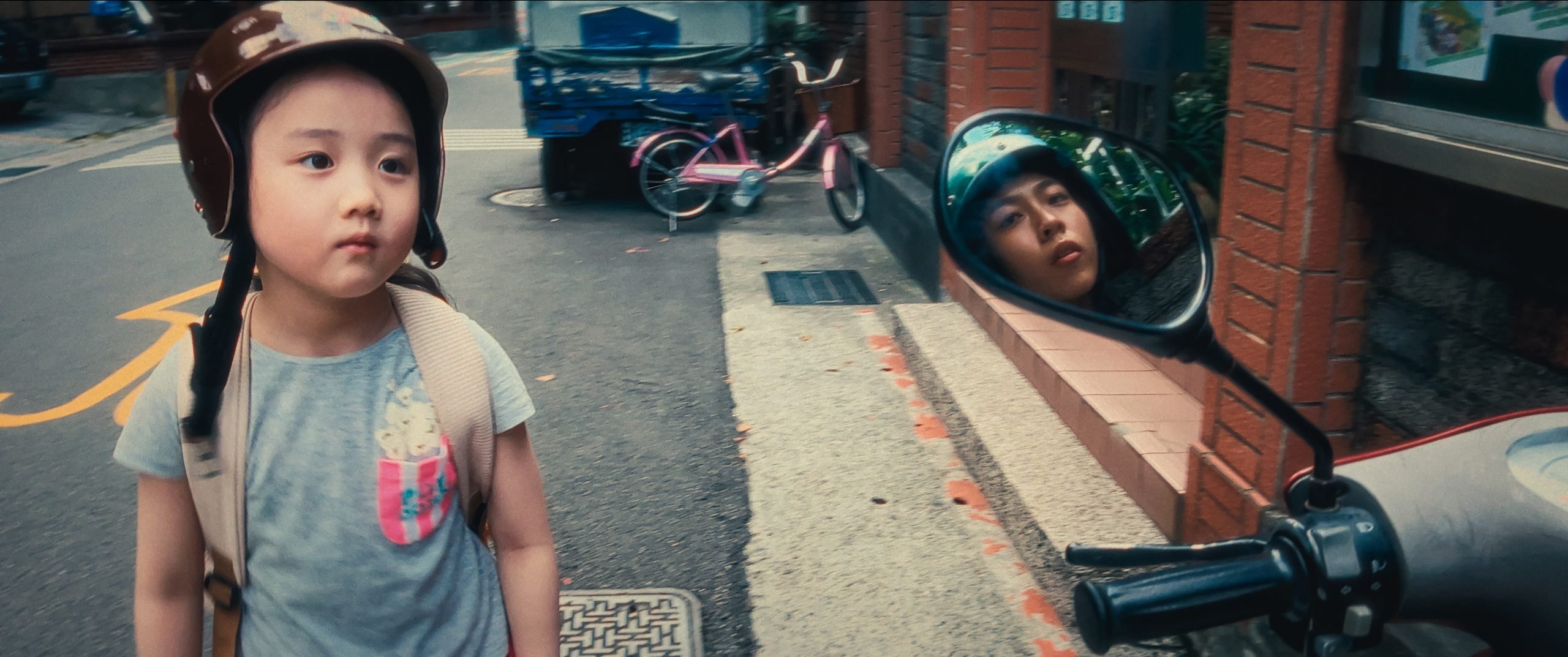 A young child wearing a helmet and backpack stands on a street, while another child's face appears in a scooter mirror in the foreground.