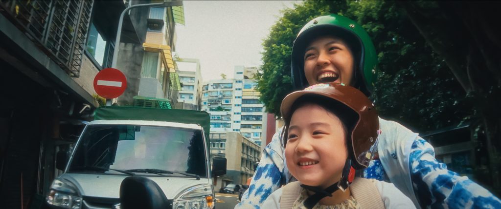 A woman and a young girl, both wearing helmets, ride a scooter on a city street lined with buildings and parked cars.
