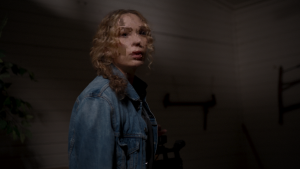 A person with curly hair wearing a denim jacket stands indoors, looking over their shoulder with a serious expression.
