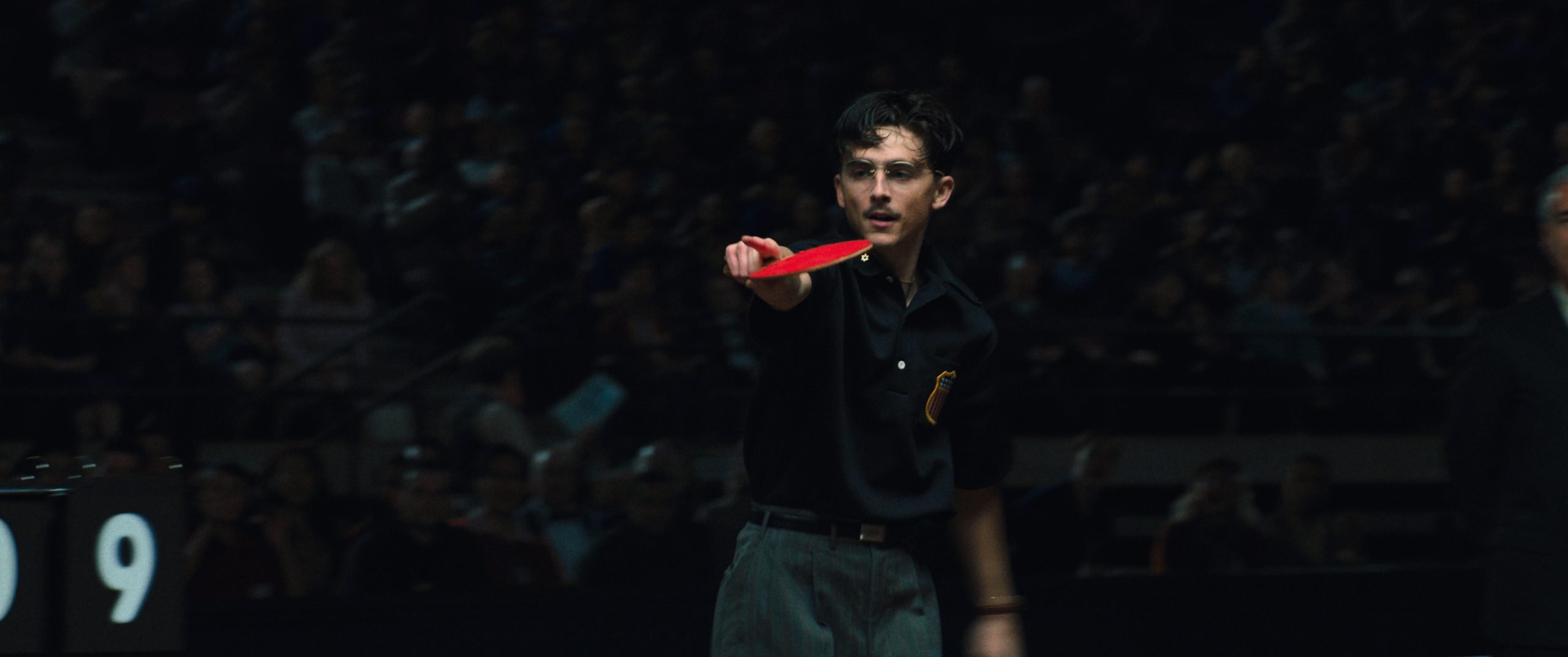 A young man in a black shirt stands in an arena, holding a red table tennis paddle and pointing, with a blurred crowd in the background.