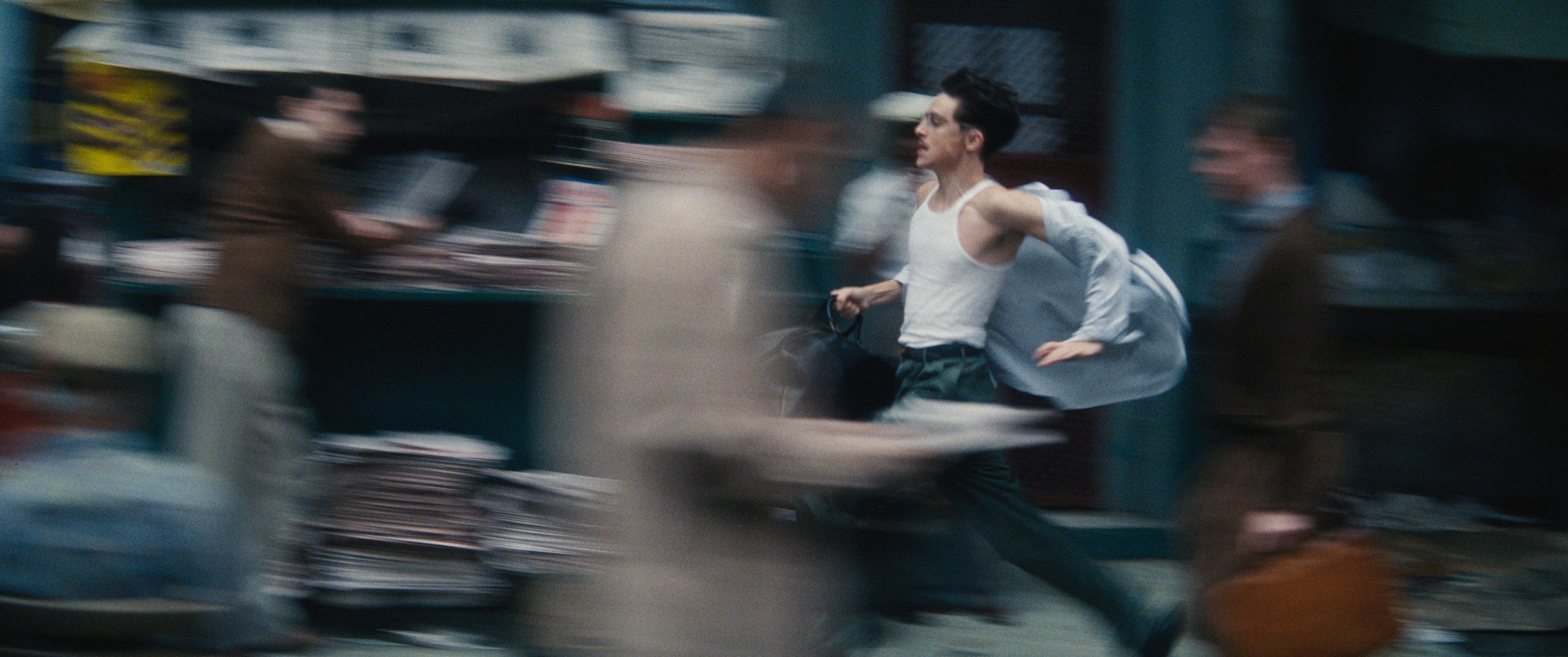 A young man in a white tank top and dark pants runs through a busy street, putting on a shirt, with blurred figures and newspapers in the background.
