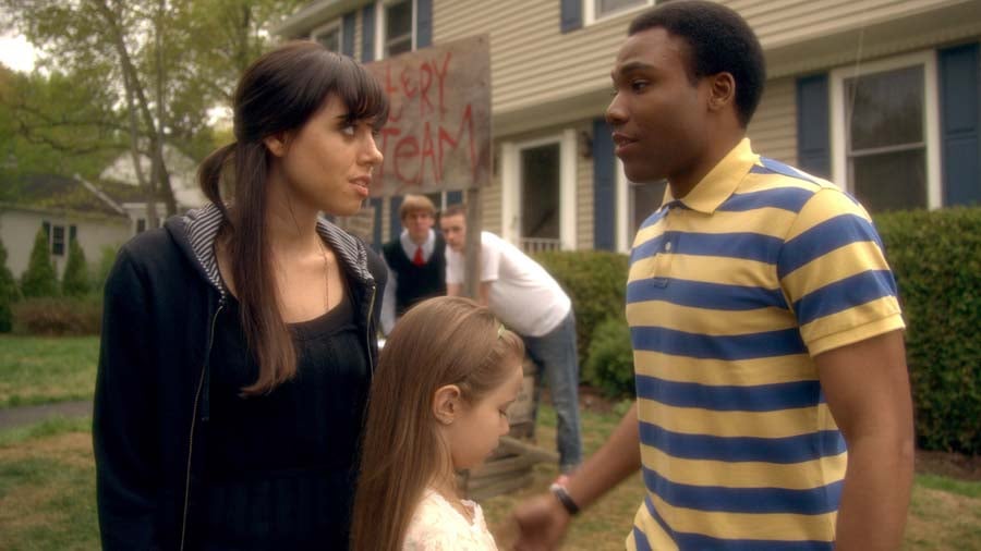 A woman, a man in a striped shirt, and a young girl stand together outside a house, with two people and a handwritten "Sorry Team" sign in the background.