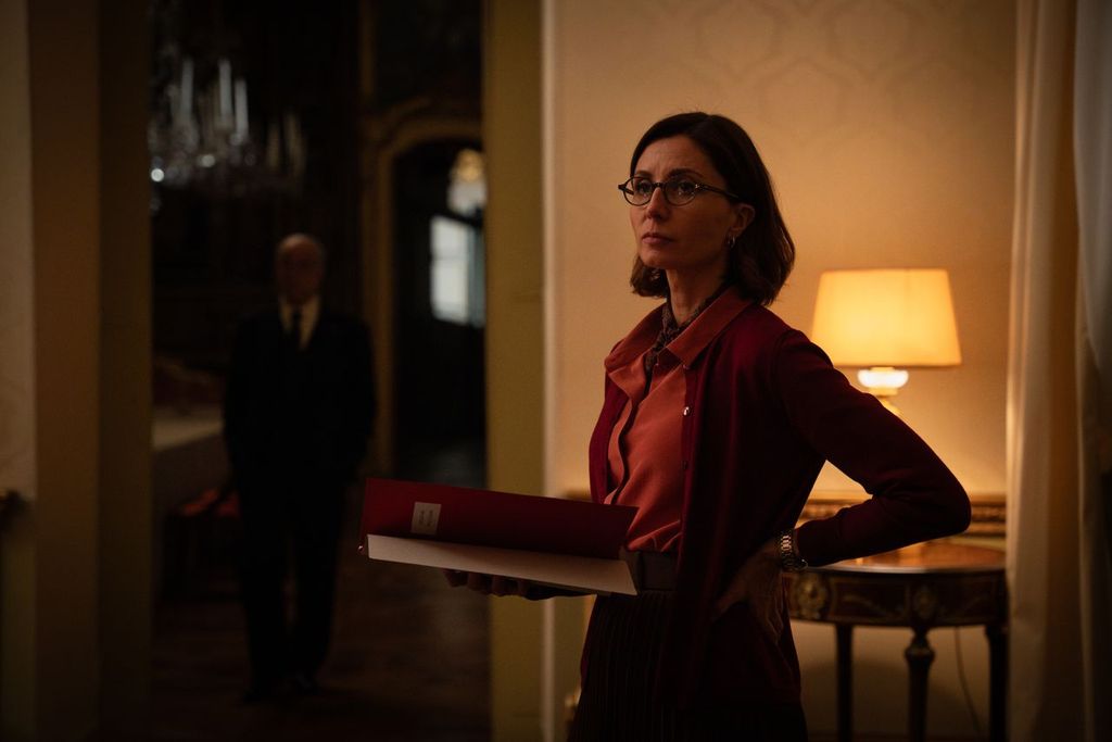 A woman in glasses holding a folder stands in a dimly lit room with a lamp on a table; a man is blurred in the background.