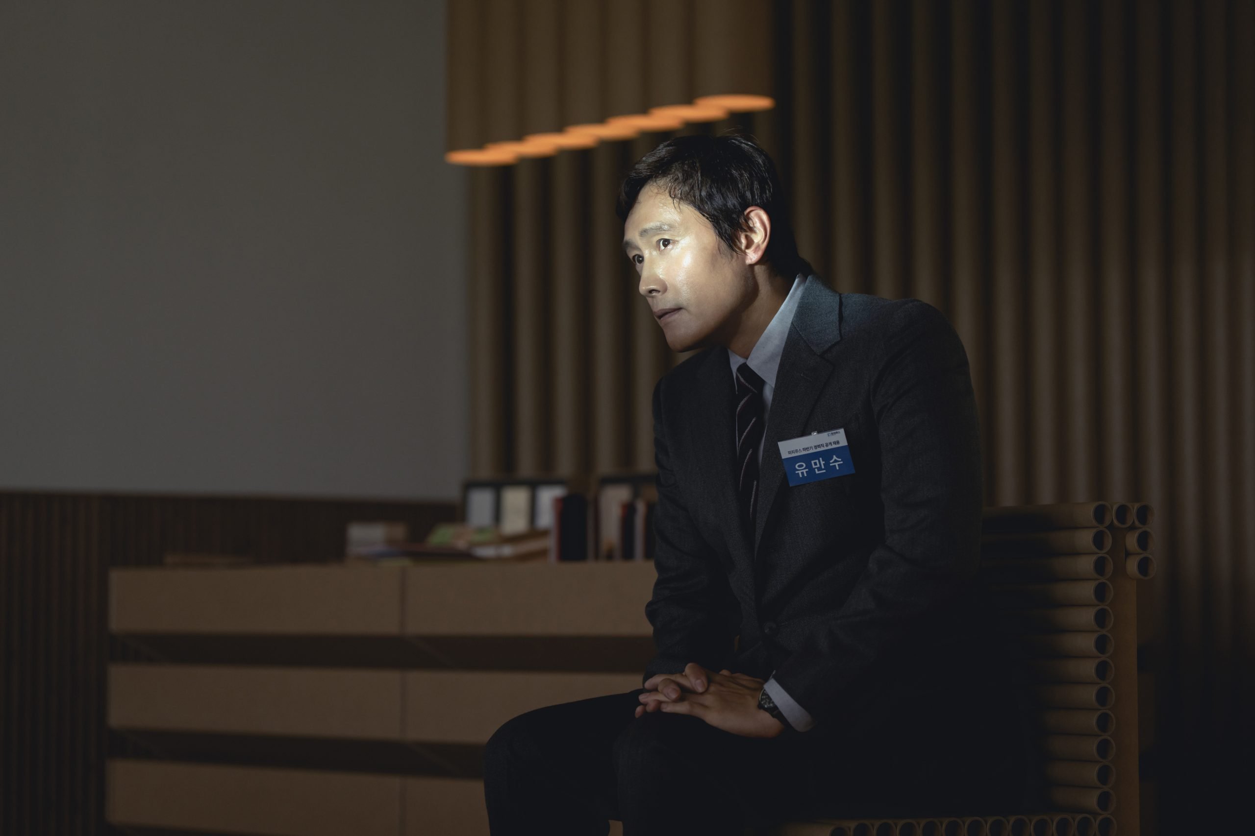 A man in a suit with a name tag sits on a bench in a dimly lit office, facing forward with hands clasped.