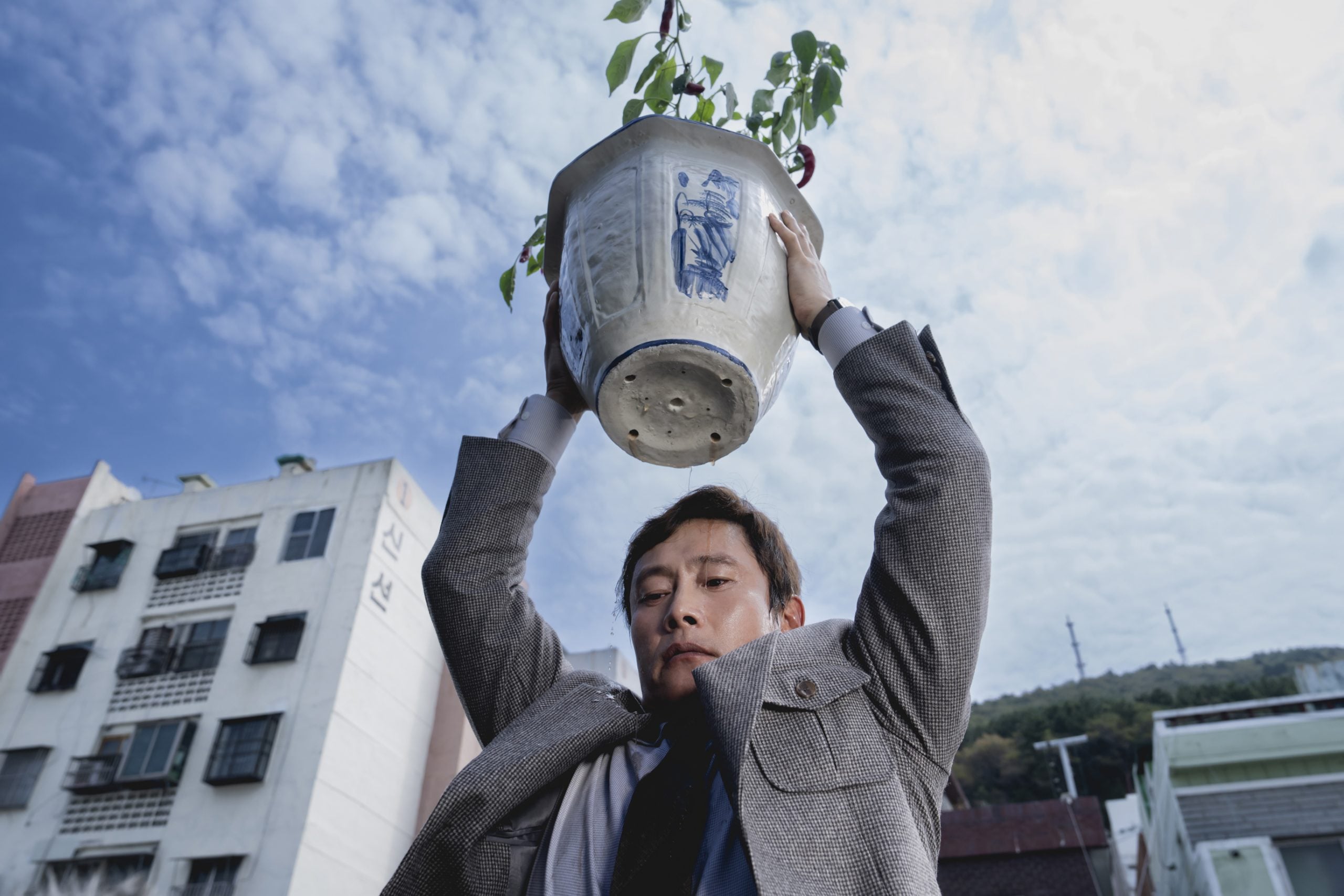 A man in a suit lifts a large ceramic plant pot above his head outdoors, with apartment buildings and a cloudy sky in the background.