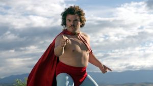A man in a red cape and leggings poses outdoors with a serious expression, pointing forward with one hand against a cloudy sky backdrop.