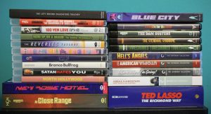 A stack of DVD and Blu-ray cases arranged in two columns, including titles like "Ted Lasso," "Hell’s Angels," and "New Rose Hotel," against a teal background.