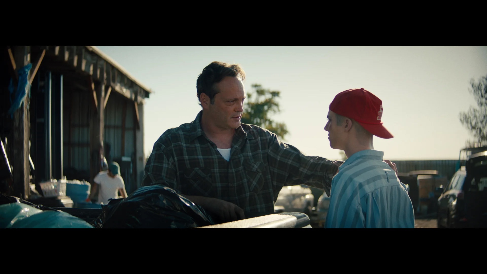 A man in a plaid shirt places his hand on the shoulder of a younger person in a red cap, talking near a structure with bags and barrels in an outdoor setting.