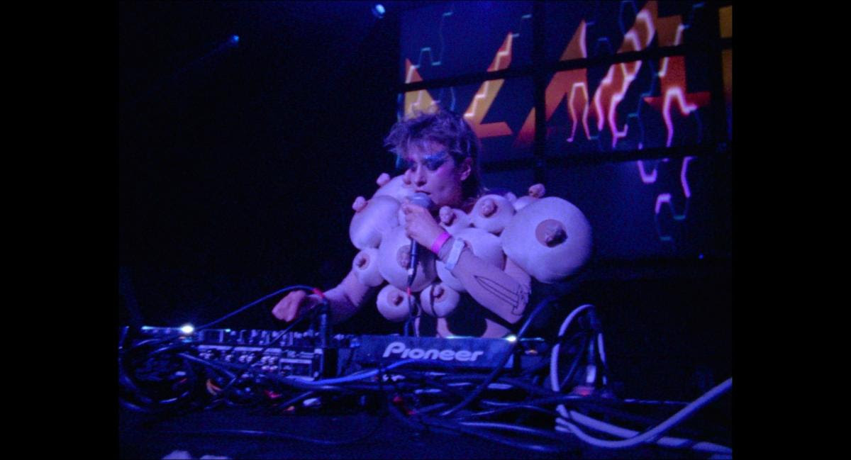 A DJ wearing a costume with multiple padded lumps performs at a Pioneer DJ setup, with colorful abstract visuals displayed on screens in the background.