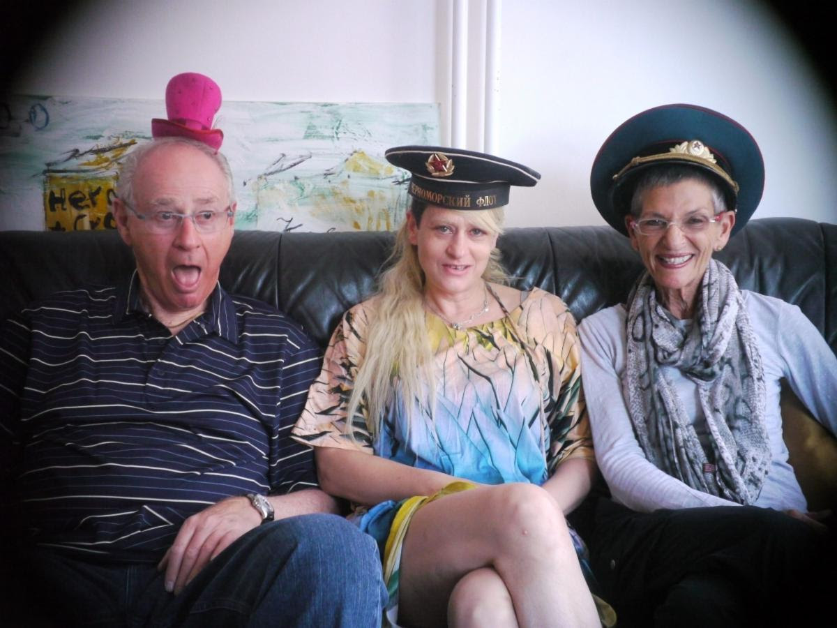 Three adults sit on a couch; the man on the left wears a small pink hat, the two women wear military-style hats, all smiling or making faces.