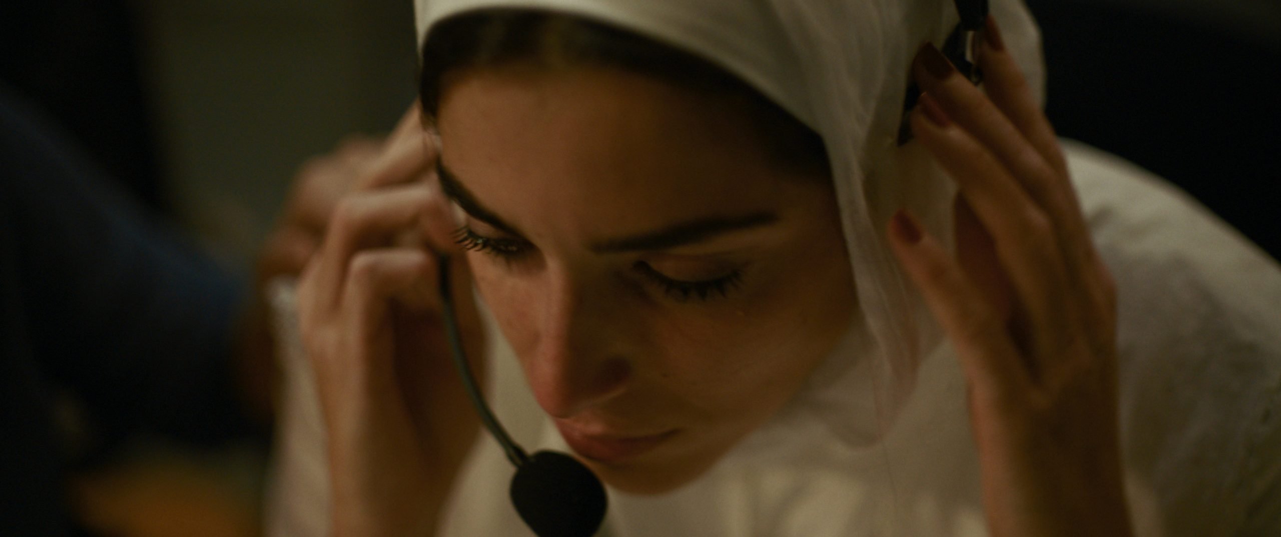 A woman wearing a white headscarf adjusts a headset microphone, looking down with a focused expression.