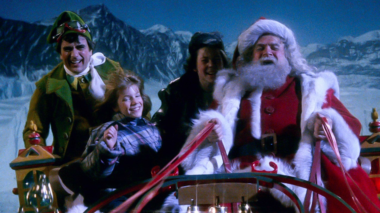 Santa Claus, two children, and an elf ride a sleigh in a snowy landscape with mountains in the background.